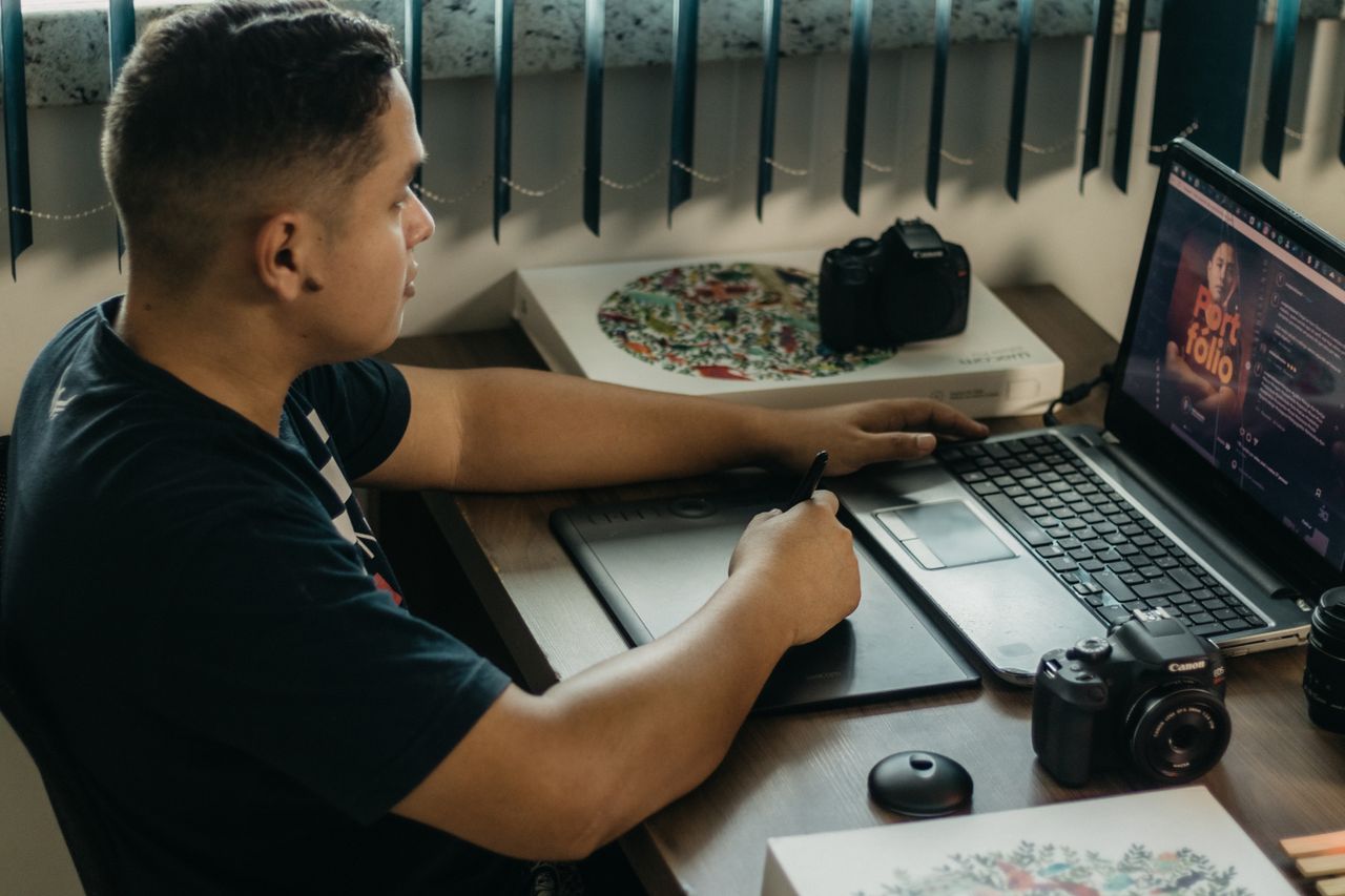 Man editing photos on a laptop using a graphics tablet, set in an indoor workspace with camera equipment.