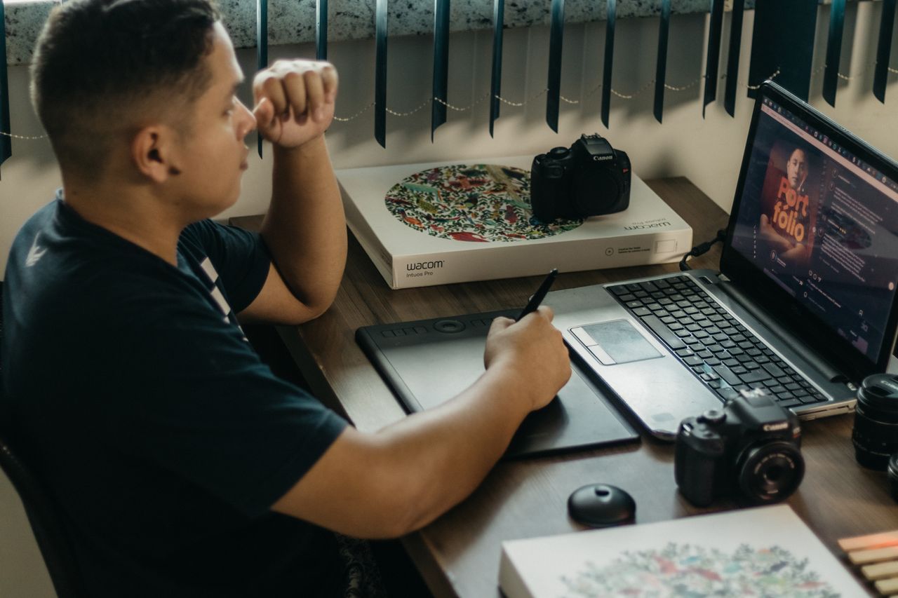 About A photographer working at a creative setup with a laptop and Wacom tablet, focused on editing photos indoors.