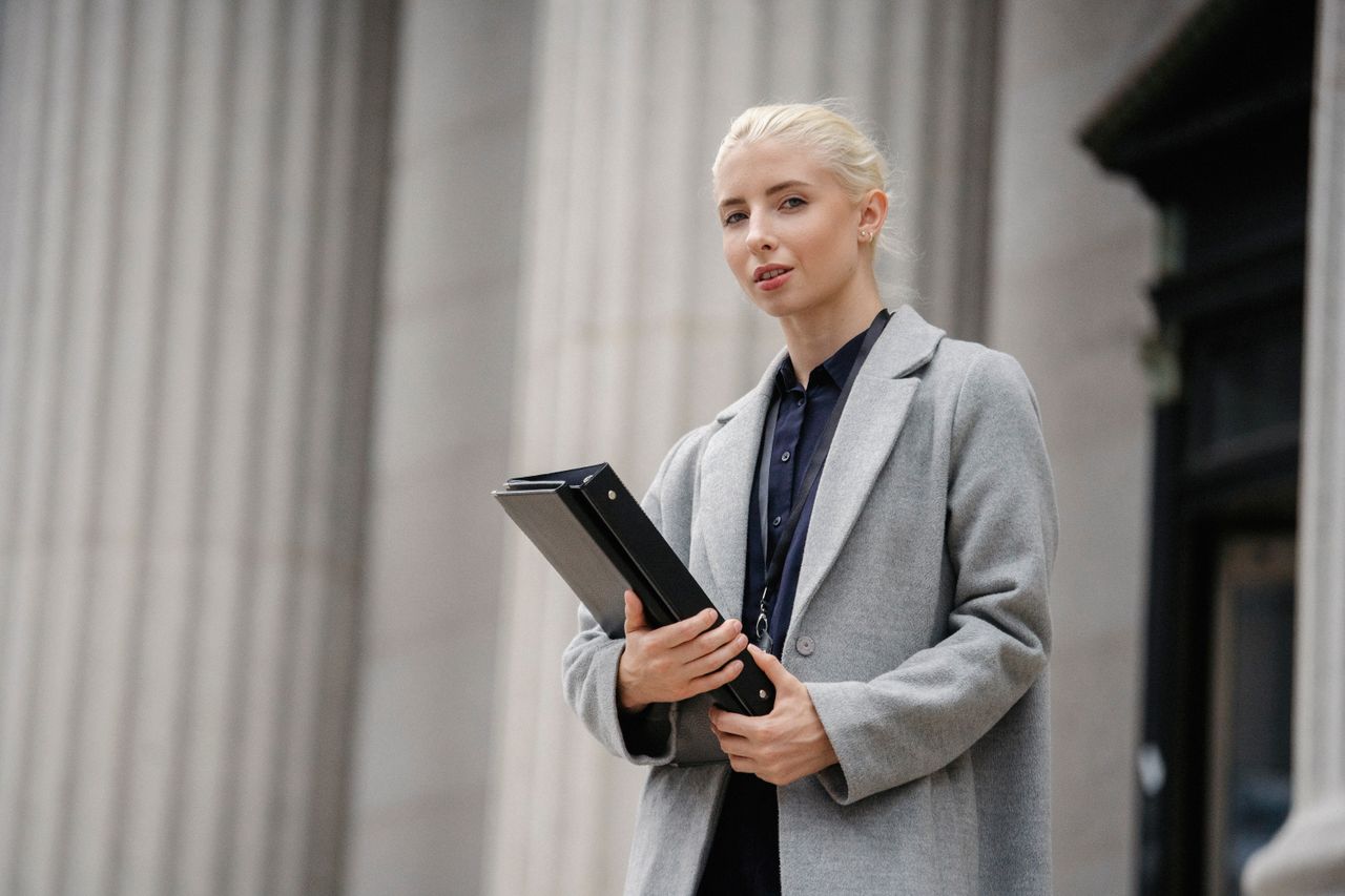 Services Calm young businesswoman in gray coat with black folder in hands standing outside stone building columns and looking at camera