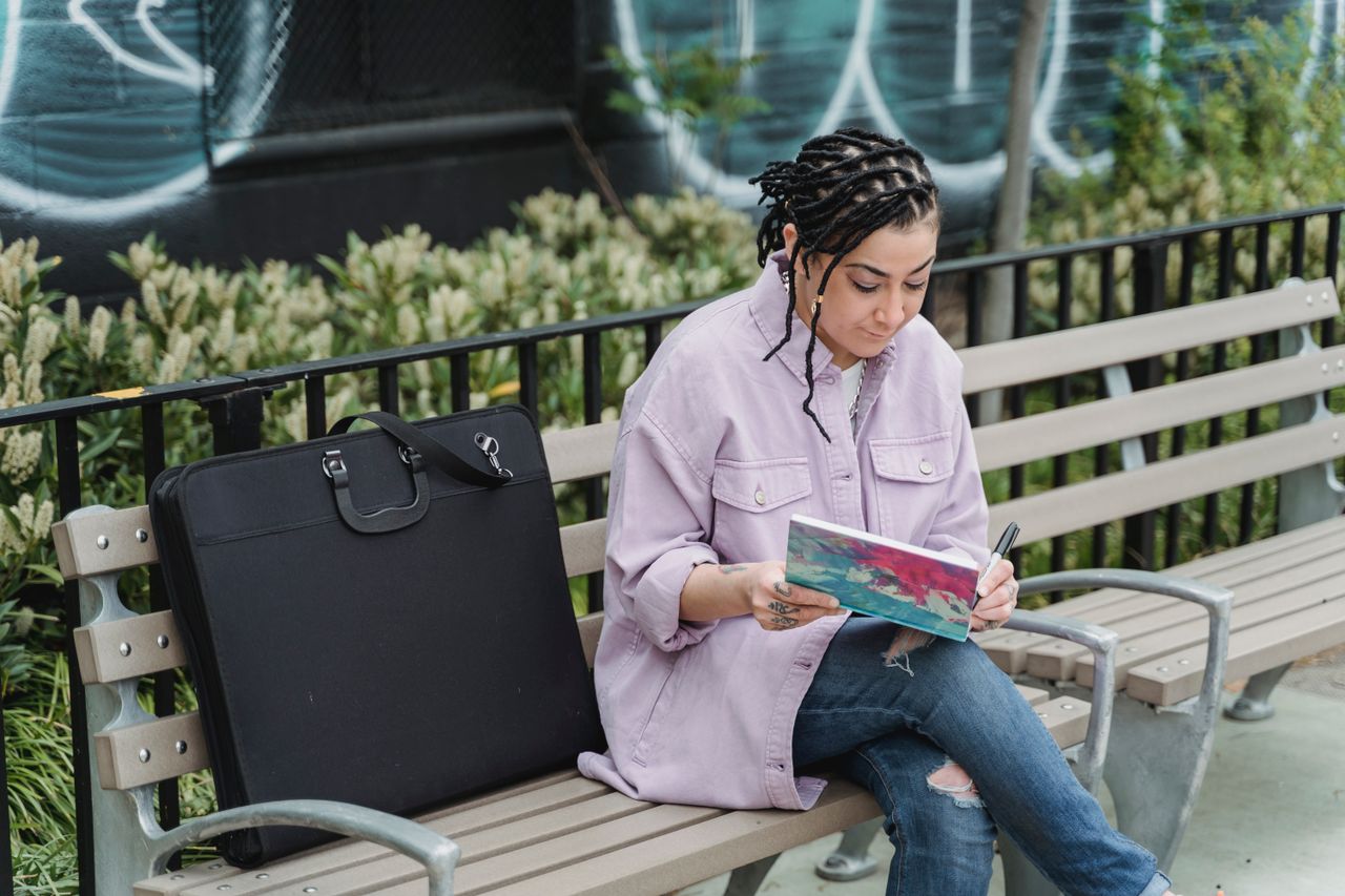 Concentrated ethnic female artist in casual clothes sitting on bench in park and drawing in sketchbook